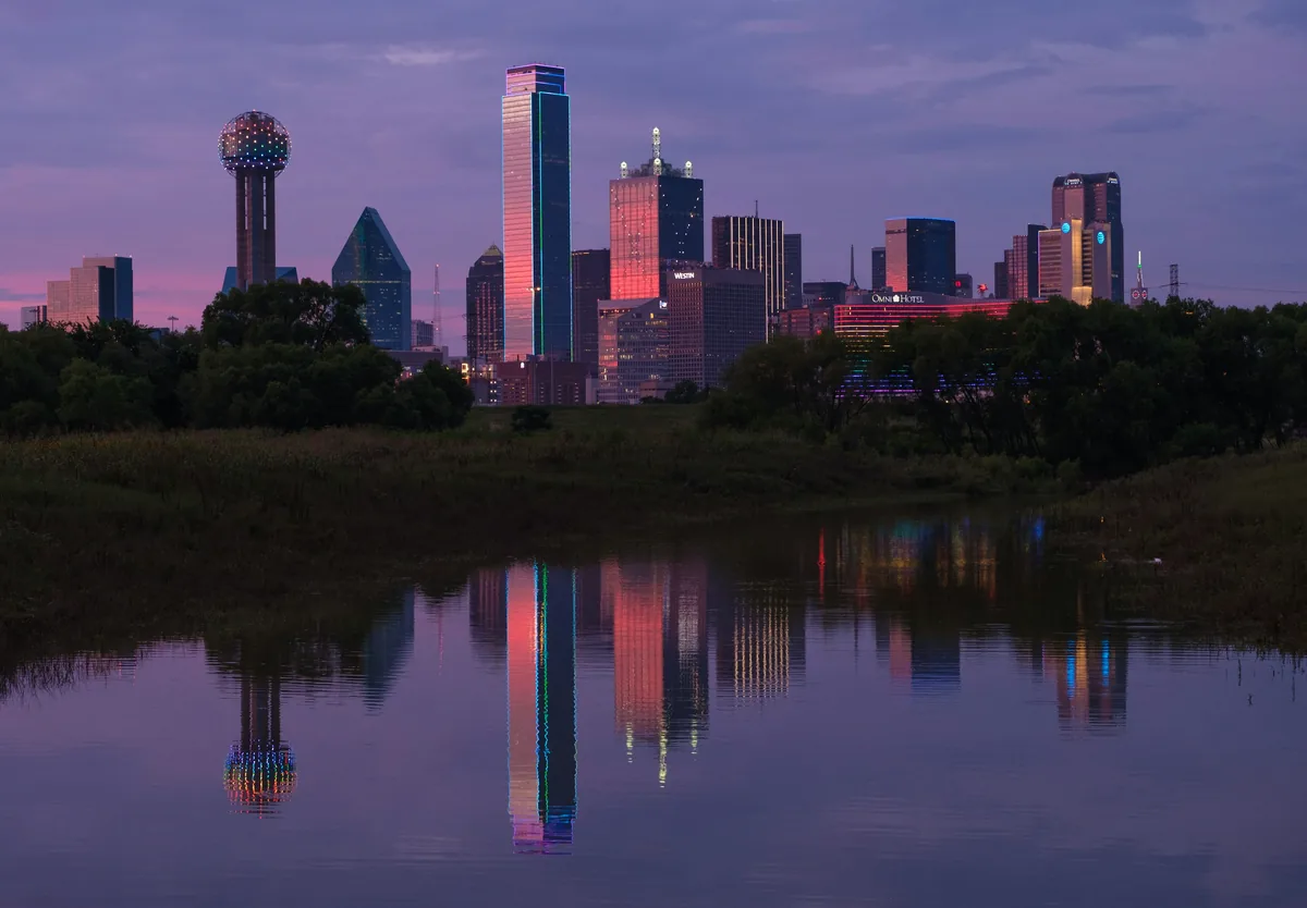 A city skyline is reflected in the still water of a pond