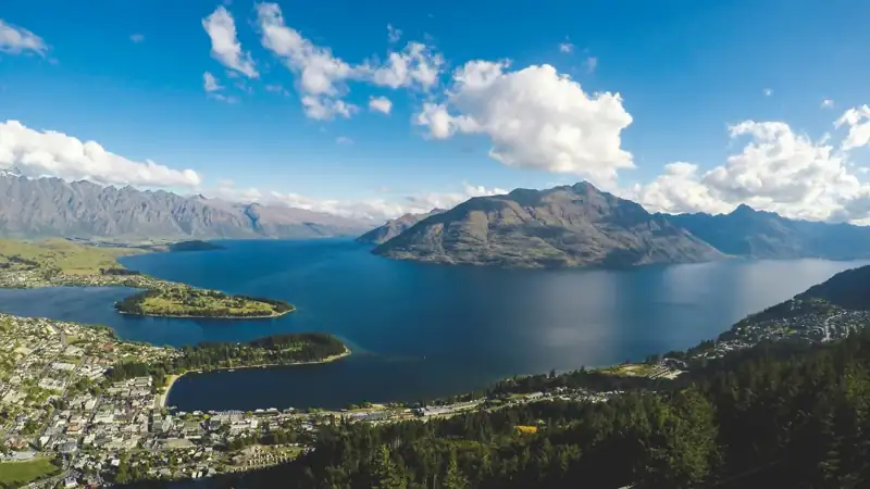 Buildings near body of water and mountains under clear blue sky and white clouds at daytime